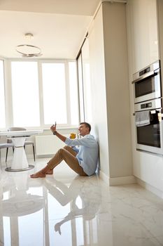 Caucasian hadsome young man in blue shirt is sitting on the floor and chilling at modern kitchen