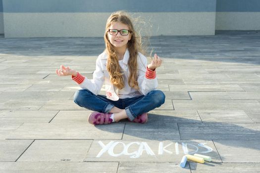 Little girl in lotus position, practicing yoga. On the asphalt text yoga kids, written by the child colored crayons
