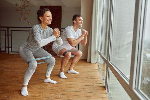 Cheerful sporty man and woman are doing squats with resistance bands before large window indoors