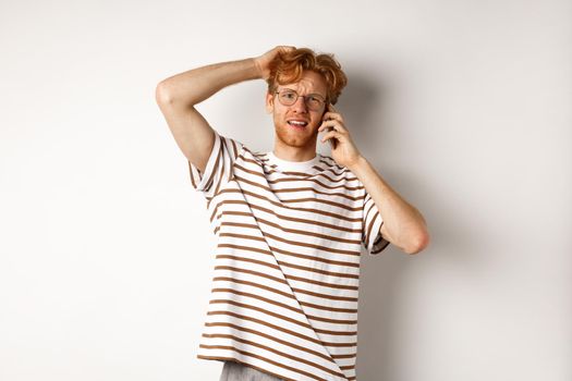 Puzzled redhead guy talking on phone, scratching head and looking confused or indecisive, standing over white background.