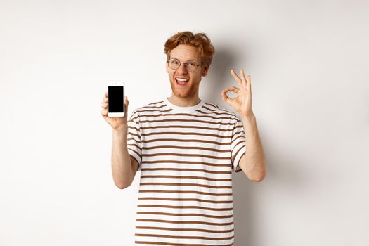 Technology and e-commerce concept. Young man with red hair showing okay sign and blank smartphone screen, praising awesome app, standing over white background.