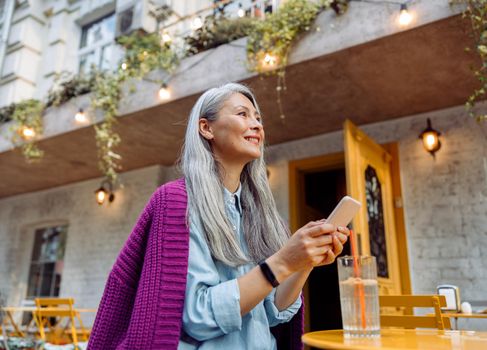 Positive senior Asian lady holds smartphone sitting at small yellow table on outdoors cafe terrace on autumn day side view