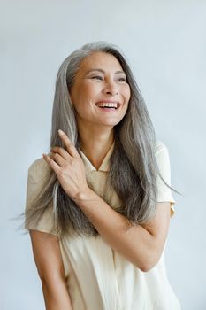 Positive middle aged Asian woman touches natural hoary hair poses for camera on light grey background in studio. Mature beauty lifestyle