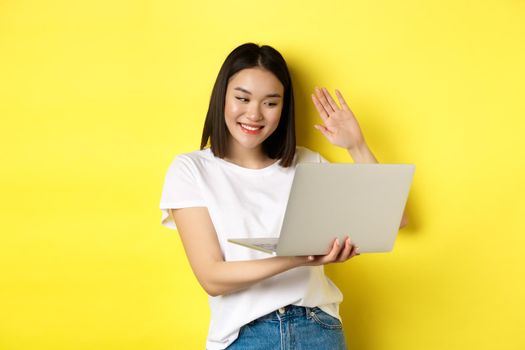 Happy asian woman video chat on laptop, waiving hand at computer camera and saying hello, standing over yellow background.