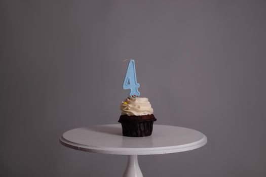 tasty chocolate cupcake with white topping cream and candle in shape of number four 4. on a white stand on a gray background. sweets, bakery. party food. dessert.