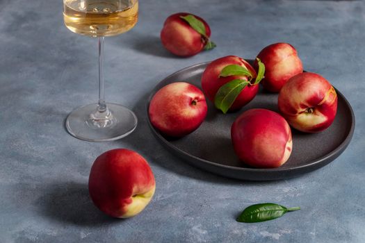 Several ripe peaches or nectarines on a black ceramic plate and a glass of white wine sit on a blue plaster-textured surface. Selective focus.