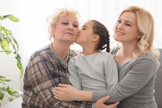 Three generations of women. Beautiful woman and teenage girl are kissing their granny while sitting on couch at home