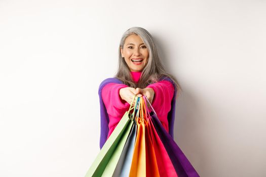 Fashionable asian senior woman going on shopping, extending hands with paper bags, smiling satisfied at camera, standing over white background.