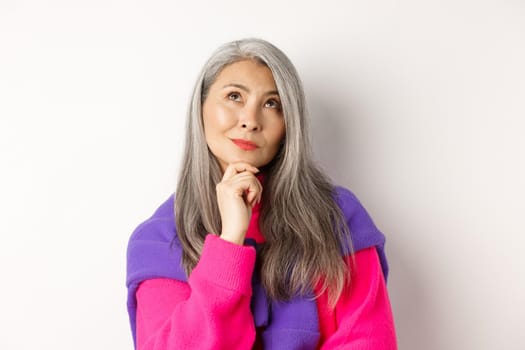 Close up of thoughtful asian female with grey hair, looking up and thinking, making decision, standing over white background.