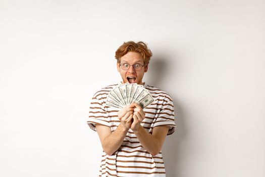 Lucky young man with red hair showing dollars, winning money and screaming of happiness, holding prize cash, standing over white background.