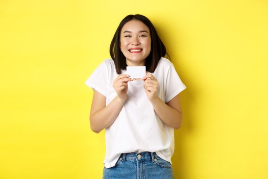 Young asian woman in casual white t-shirt going on shopping, showing plastic credit card and smiling excited, standing over yellow background.