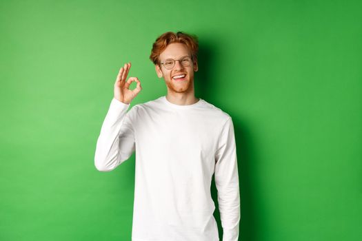 Happy male student with red hair, wearing glasses, showing okay sign in approval and smiling satisfied, standing over green background.