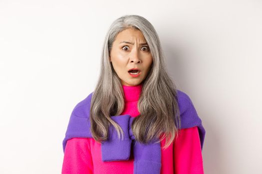 Close-up of shocked asian senior woman gasping, staring with confused face, looking with cringe, standing against white background.