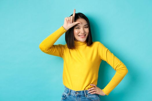 Sassy asian girl mocking lost team, showing loser sign on forehead and smiling pleased, being a winner, standing over blue background.