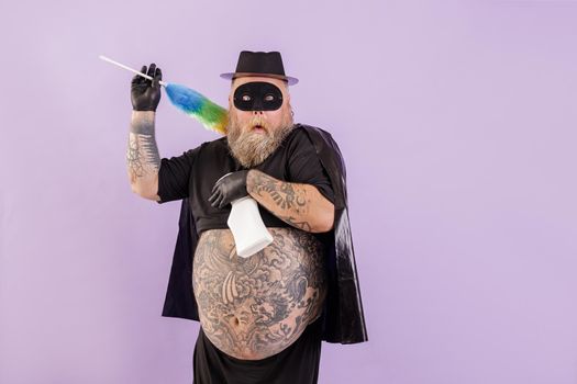 Funny scared middle aged man with overweight in hero costume with fluffy dust brush and detergent bottle poses on purple background in studio