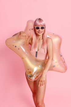 Cheerful woman with pink hair in swimsuit and sunglasses with inflatable star for swimming posing isolated over pink background. Summer vacation, fun concept