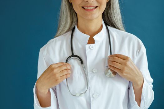 Smiling mature female doctor in white robe with stethoscope stands on blue background in studio closeup. Professional medical staff