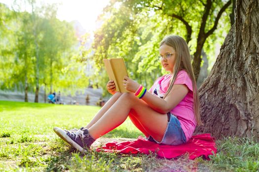 Girl child in glasses reading book in the park, on the grass near the tree