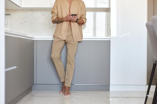 Hadsome smiling man is chilling on the minimalistic kitchen with smartphone