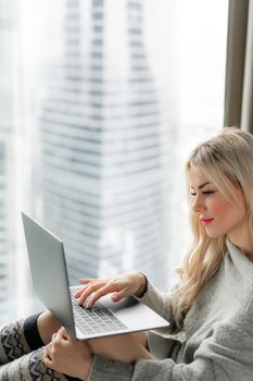 Beautiful blonde woman lying on the bed with gray aluminum laptop. Girl smiles, good mood. Blogging, browsing internet, chatting. In a warm cozy sweater and wool socks. Happy winter concept