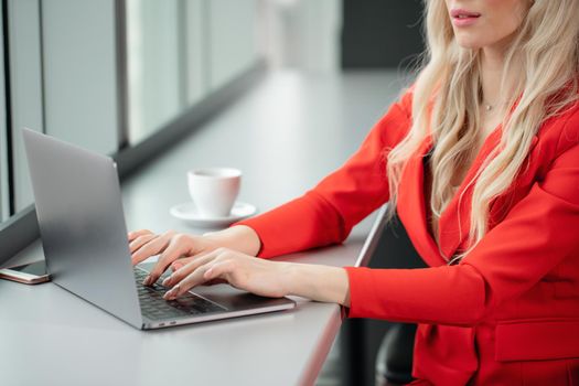 Woman in red coral business suit. Young blonde businesswoman with modern gray laptop in the office in high floor in front of the window. Girl drinking coffee.