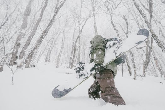 Unrecognizable freerider young man with snowboard walking in winter forest among snowdrifts