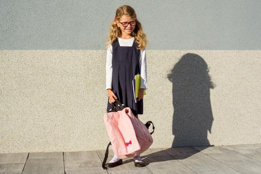 An elementary school student with notebooks in his hand. A little schoolgirl with a backpack near the building in the open air.