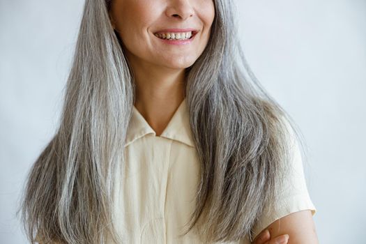 Middle aged woman with long silver hair smiles on light grey background in studio closeup. Mature beauty lifestyle