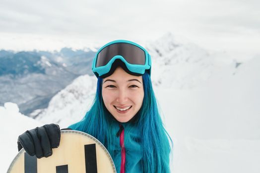 Happy young woman with blue hair standing with snowboard on background of snowy mountains, looking at camera