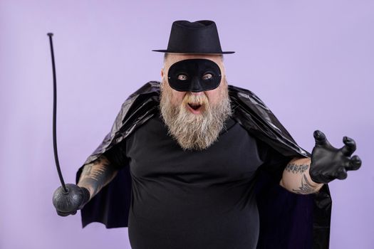 Funny mature bearded man with overweight wearing Zorro suit with cape, mask and toy epee poses for camera on purple background in studio