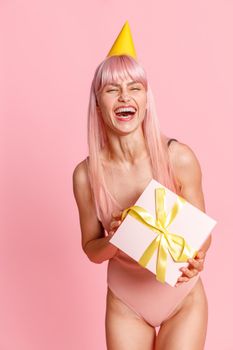 Excited young woman with pink hair wearing swimsuit and party hat holding cute gift box, standing isolated over pink background. Holiday, birthday concept