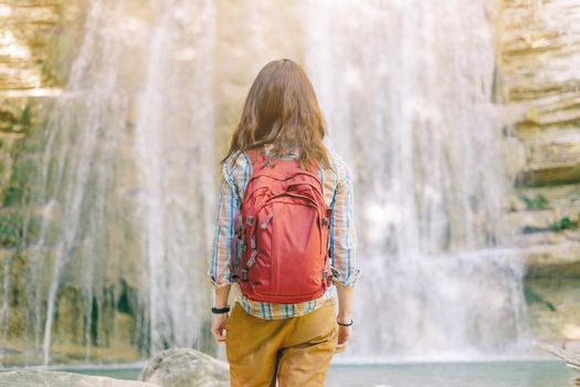 Unrecognizable hiker woman with backpack standing in front of waterfall, rear view.