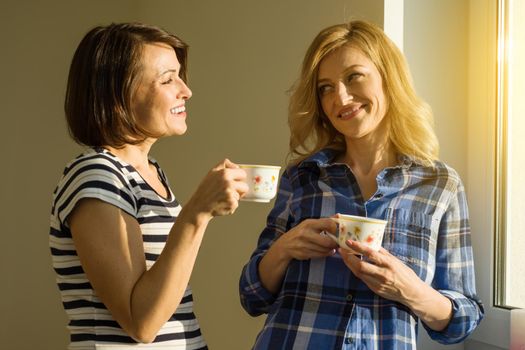 Beautiful adult woman holding hot cups of coffee drinking, standing near the window and looking at each other sharing funny things, enjoying sisters talking time