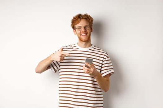 Technology and e-commerce concept. Young man with red messy hair, wearing glasses and t-shirt, pointing finger at smartphone and smiling satisfied, white background.