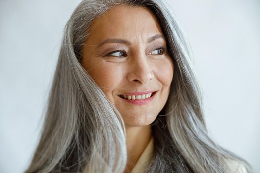 Attractive middle aged Asian lady with loose grey hair poses for camera on light background in studio closeup. Mature beauty lifestyle