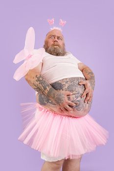 Surprised bearded plump male person in carnival fairy costume with pink wings holds large tummy on purple background in studio