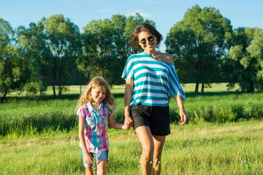 Woman with daughter child walking in nature holding hands. Background blue sky, green grass, small river.