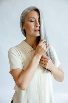 Languid middle aged Asian woman in blouse strokes lock of silver hair standing on light grey background in studio. Mature beauty lifestyle