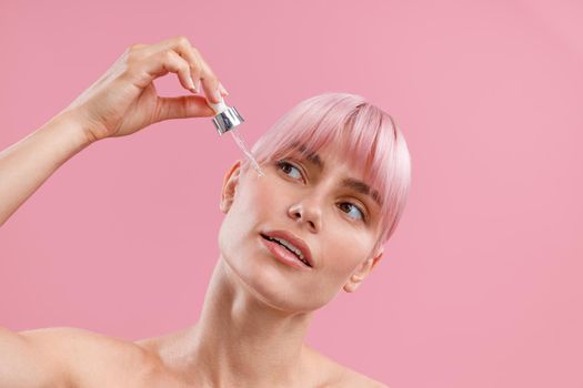 Portrait of young woman with pink hair applying serum or hyaluronic acid on her facial skin using dropper, posing isolated over pink background. Beauty, cosmetics and skincare concept