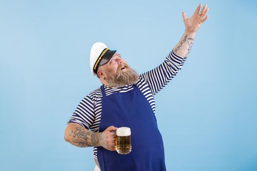 Positive middle aged bearded man with overweight in sailor costume with apron holds mug of beer raising up hand on light blue background in studio