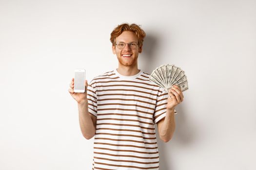 Smiling young redhead man in glasses showing smartphone blank screen and money, standing over white background.