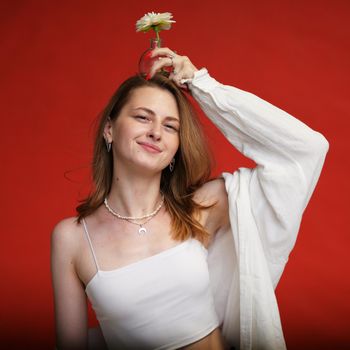 Caucasian smiling girl wearing white clothes posing with flower in studio on red background