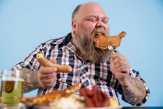 Expressive bearded person with overweight eats smoked chicken leg holding sausage at table with greasy food on light blue background in studio