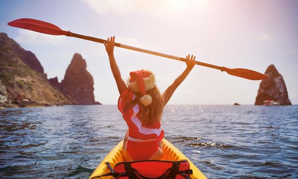 Young brunette woman in red swimsuit and Santa hat, swimming on kayak around basalt rocks like in Iceland. Back view. Christmas and travel concept