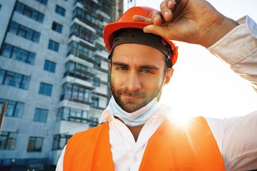 Portrait of man builder in workwear and hardhat wearing medical mask, close up photo