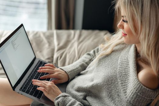 Beautiful blonde woman lying on the bed with gray aluminum laptop. Girl smiles, good mood. Blogging, browsing internet, chatting. In a warm cozy sweater and wool socks. Happy winter concept