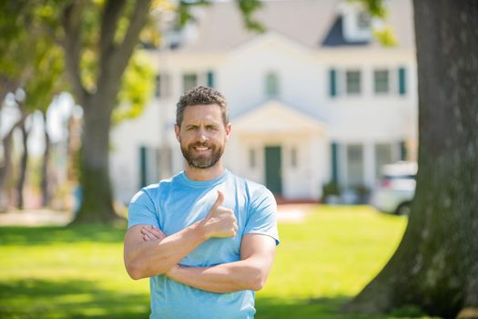 happy bearded man standing showing thumb upon house background, broker.