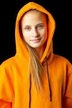 Shot of beautiful stylish teenage girl. Portrait of attractive teenager wearing warm orange hoodie putting the hood on. Beautiful brown eyed girl posing against black background