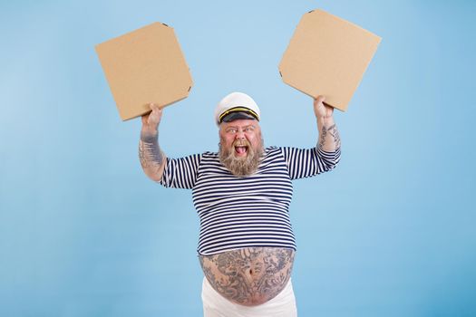 Joyful middle aged bearded man with overweight in sailor costume holds blank cardboard boxes of pizza screaming on light blue background in studio