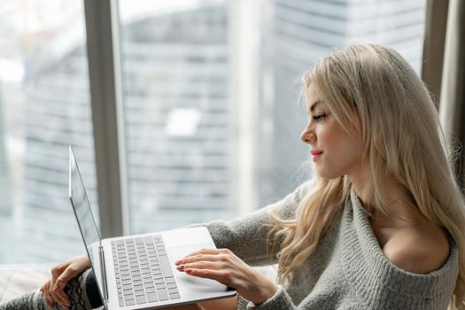 Beautiful blonde woman lying on the bed with gray aluminum laptop. Girl smiles, good mood. Blogging, browsing internet, chatting. In a warm cozy sweater and wool socks. Happy winter concept